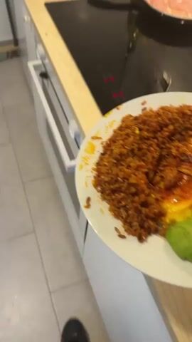 Here's a detailed description of the video:

The video shows a plate of food and a pan of cooking meat in a kitchen setting. The plate contains a portion of brown fried rice, sliced avocado, and what appear to be fried plantains. On the stovetop next to the plate, there is a black frying pan containing thinly sliced raw chicken with some garlic pieces, cooking in oil. The stovetop is black, and other items visible include a bottle of oil and some dish towels. The kitchen has white cabinets and a tiled floor. The camera moves slightly, focusing on different elements of the scene.