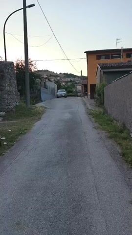 This video captures a serene, rural scene in Campochiaro, Italy, at dusk. The camera pans along a narrow, paved road, revealing the quiet charm of a small Italian village. 

The scene opens with a view of the road ahead, with a silver car parked in the distance. To the left, a weathered stone wall and lush greenery create a rustic foreground, contrasting with a modern street lamp and overhead wires. On the right, the warm tones of an orange building stand out against the twilight sky.

As the camera moves forward, it passes an old, dilapidated building on the left. This structure, with its stone foundation, rough stucco walls, and a boarded-up window, exudes a sense of history and abandonment. A wooden fence with a bright yellow warning sign adds a touch of visual interest. The sign reads "PERICOLO - BAGNI" which translates to "DANGER - BATHROOMS", suggesting the building might have once housed communal facilities or perhaps a more private, but now unsafe, restroom.

The camera continues its slow progress, offering glimpses of more houses and structures in the background, nestled against a gentle hillside. The sky transitions from a pale blue to a soft orange hue, indicating the setting sun. The overall atmosphere is one of peacefulness and quietude, characteristic of a rural Italian village at the end of the day. The lighting and the stillness of the scene evoke a sense of calm and introspection.