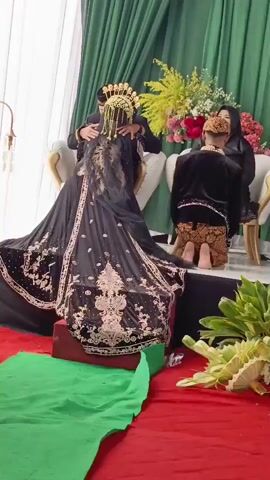 The video shows a wedding ceremony with several people. In the foreground, a person in a black wedding dress with gold embellishments and a traditional headpiece is being embraced by another person in a black outfit. To the right, another person, presumably the groom, is kneeling in a black outfit with traditional patterned pants, facing away from the camera. They are wearing a head covering. Behind them, another person in a dark outfit and head covering is seated. The background features a green curtain and a floral arrangement with red and pink flowers. There are also white chairs and a gold-colored lamp fixture visible. The floor is covered with red and green fabric. The lighting suggests an indoor setting. The audio contains speech, possibly from the ceremony.