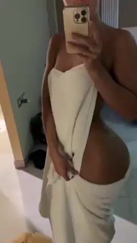 A slender woman stands in a bathroom mirror, holding her phone and taking a selfie. She is partially covered with a white towel. Her body is tanned and toned. The background includes a white bed, electrical outlets, a small black fan, and light-colored floor tiles. The audio has male speech.