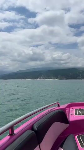 The video shows a pink speedboat moving on a body of water. The boat's interior is predominantly pink with black and light pink seating. The camera faces forward, showing a wide expanse of water ahead with a distant shoreline featuring green hills or mountains. The sky is partly cloudy with patches of blue visible. There are no visible people on board. The speedboat is moving at a steady pace, creating small waves.