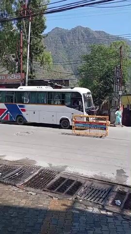 The scene is a street in a town or city surrounded by mountains. A large white bus with a blue and red design is parked in the middle of the road, partially obstructed by a yellow and black barrier. A person dressed in light blue clothing walks on the sidewalk in the distance. Another person wearing a red shirt and gray pants walks across the street in front of the bus. Buildings line the street, including one with a sign that reads "Adventure India." The sky is blue and the sun is shining.
