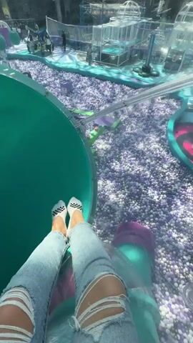 This video captures a first-person perspective of someone going down a brightly colored slide. The subject is wearing ripped blue jeans and white socks with a black checkered pattern.

The slide appears to be part of an indoor play area, indicated by the large ball pit filled with purple and white balls visible at the beginning and end of the footage. There are also glimpses of other play structures and what looks like a bright, artificial environment. The slide itself transitions through different colors, starting with a teal tube, then a bright pink tube, and finally returning to a teal section with the ball pit below.

The activity is clearly recreational, with the subject experiencing a thrilling ride down the slide. The rapid color changes and the blur of motion suggest speed and excitement. The lighting appears to be bright and artificial, typical of an indoor amusement setting, making it difficult to determine the time of day or weather. The overall mood conveyed is one of fun and exhilaration. There is no visible text in the media file. The location context provided indicates "sovkhoza imeni Lenina, Russia."