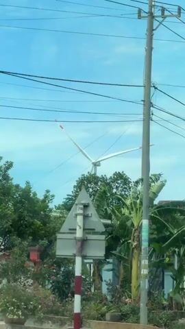 A short video clip shows a view from a moving vehicle looking towards a rural landscape. Banana trees with large green leaves dominate the foreground. In the background, a tall, white wind turbine with two visible blades stands against a bright blue sky with scattered clouds. Power lines are strung across the sky. There are also some buildings, including one with a corrugated metal roof and a grey brick structure with a door. The trees and leaves are visibly swaying, indicating a windy day. The overall scene depicts a somewhat underdeveloped area with both natural elements and infrastructure.