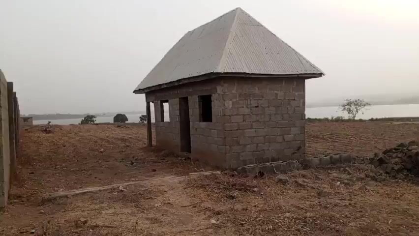 The video shows a landscape with a small unfinished brick building to the left. It has a grey corrugated metal roof. The land is mostly dry and brown with patches of vegetation. A lake is visible in the distance, with hills on the far side. The sky is overcast, with the sun barely visible through the haze. A dirt road runs along the right side of the frame.