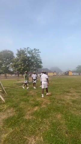 The video shows a group of young men in white uniforms on a grassy field under a hazy sky. They appear to be engaged in a training exercise or recreational activity involving a soccer ball. One man is kicking the ball, while others observe or participate. The environment includes trees, buildings in the background, and some exercise equipment. The overall scene suggests an outdoor training camp or a similar organized setting.