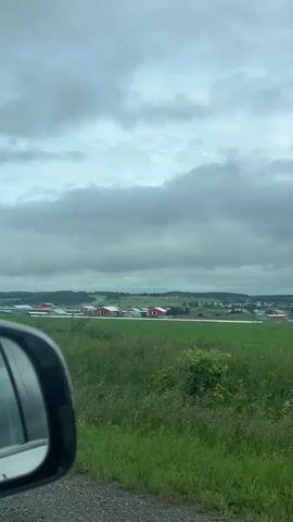 A car drives past a large green field with a line of red-roofed buildings in the distance. The sky is cloudy and overcast. The side mirror of the car is visible in the foreground.