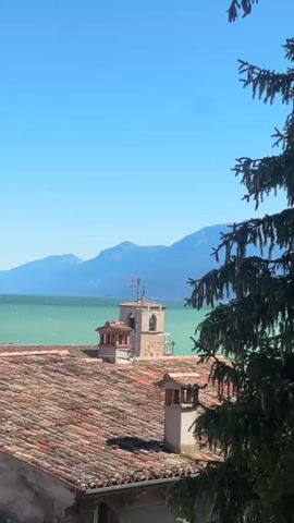The video captures a scenic view of a body of water, likely a lake, with mountains in the background under a clear blue sky. In the foreground, there are red-tiled roofs of buildings, some with chimneys. A tall tower with a few small domes and a smaller clock tower are visible among the rooftops. A large tree with green needles partially obstructs the view on the right side. The water has a turquoise hue, and there are faint ripples on its surface, suggesting a gentle breeze. Distant shoreline features buildings and more vegetation. A satellite dish and a multi-pronged antenna are also visible on one of the roofs. The overall impression is that of a sunny day in a picturesque, possibly Mediterranean, coastal town.