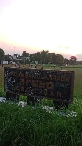 A sign is in the foreground, partially obscured by tall green grass. The sign is weathered and has dark lettering on a lighter background, with the visible text including "LAPANGAN OLAHRAGA" and "KEBOHARAN". Beyond the sign is a large, grassy field. In the distance, there are several buildings and trees. Several people, likely playing a sport, are visible on the field. The sky is a pale orange and pink, suggesting either sunrise or sunset. There are also light poles on the field.