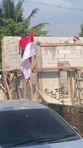 The video shows an outdoor scene with a partially constructed building made of concrete blocks. A wooden scaffolding surrounds the building. A person wearing a red shirt and a head covering is standing on the scaffolding, holding a flag pole with the Indonesian flag attached. The flag is red and white. Another person, wearing a red and white striped shirt and a red and white hat, is visible on top of the building. There are trees in the background and a car in the foreground. The sky is blue with some clouds.