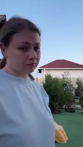 A woman with brown hair pulled back in a ponytail, wearing a light blue t-shirt, stands in front of a playground. She is looking directly at the camera with a slight smirk. Behind her is a wooden playground structure with a yellow slide and various toys scattered on the ground. The background shows a clear sky with a few distant buildings. The scene appears to be outdoors, possibly in a backyard, during daylight.