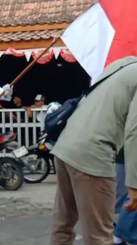 This video captures a procession featuring several individuals dressed in white uniforms and black caps, holding flags. The person in the foreground on the left is holding a long wooden pole with a white flag attached. To their right, a person in a similar uniform is holding a green flag with text that appears to read "YAYASAN PENDIDIKAN AL-AZHAR WIROSARI GROBOGAN". Further to the right, another person in a white uniform holds a yellow flag. Behind the procession, a motorcycle is parked, and there are people visible behind a fence. The background shows a building with a tiled roof and red and white decorative trim. The overall scene appears to be an outdoor event or parade.