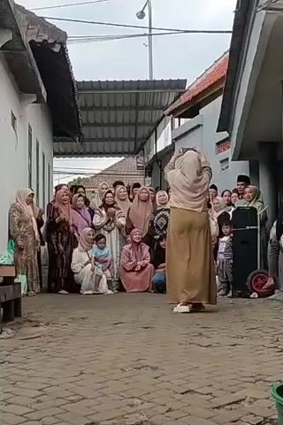 A group of people are gathered in an outdoor alleyway between two buildings, possibly for a celebration. Most of the people are women and children, dressed in traditional Islamic attire. A woman in the foreground, facing away from the camera, is holding up a phone to record or photograph the group. The alleyway is paved with bricks and has covered walkways on either side. The sky is overcast.