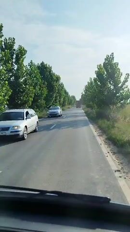 A first-person perspective video shows a car driving on a paved road. Trees line both sides of the road, casting shadows on the asphalt. The sky is clear with some clouds. A silver station wagon is visible on the left side of the road in the foreground. Further down the road, other vehicles, including what appears to be a truck, can be seen in the distance. The video captures the movement of the car through the landscape.