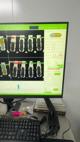 The video shows a close-up of a computer monitor displaying a quality control interface, likely for pharmaceutical production. The screen displays multiple images of capsules, with some highlighted in red, indicating potential defects. Data fields on the right side of the screen show numerical values and status indicators, including "11.66". Below the monitor, a keyboard and a desk are visible. The desk has papers, a cell phone, a roll of tape, and what appears to be a red warning light. The camera then pans down to a large stainless steel bin filled with yellow capsules, suggesting the context of the quality control is automated capsule inspection. The background is a plain white wall, typical of an industrial or laboratory setting.