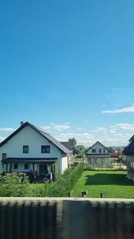 A video captures a series of houses under a bright blue sky with scattered clouds. The houses appear to be residential, with visible roofs, windows, and some with yards. Green trees and bushes are present around the properties. The camera seems to be moving, possibly from a vehicle, as indicated by the slight blur and consistent perspective shift. Some houses appear newly constructed or under renovation, with visible building materials and fencing in the yards. Cars are visible in some of the driveways. The overall scene depicts a suburban or semi-rural residential area during daylight.