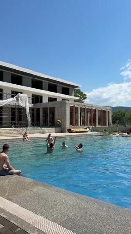 This video shows a group of young people swimming and playing in a pool on a sunny day. In the background, a building is under construction, with scaffolding and construction materials visible. The pool water is a vibrant blue, and the sky is clear with a few clouds. The people in the pool appear to be enjoying themselves, some are splashing, and others are throwing a ball. The overall atmosphere is relaxed and summery.