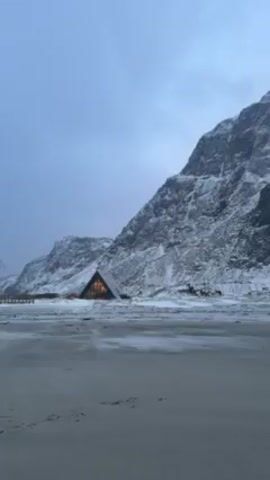 The video shows a beach scene on a cloudy day, with snow-covered mountains in the background. There is a small A-frame building lit up in the distance. The camera pans across the beach towards the ocean. A person on a surfboard can be seen sitting in the water.