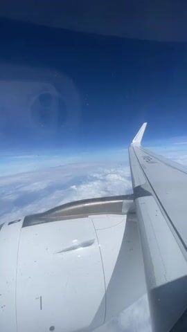 The video shows an aerial view from an airplane window. The foreground features the wing and engine of the aircraft, with the wing extending towards the right of the frame. The engine is white with metallic accents and panel lines. Below the wing and engine, a layer of white clouds stretches across the horizon. Above the clouds, a deep blue sky dominates the upper portion of the frame. There is a circular reflection on the window in the upper left quadrant. The video appears to be a static shot, with no discernible movement of people or significant action. The environment is clearly that of an airplane in flight, high above the cloud layer.