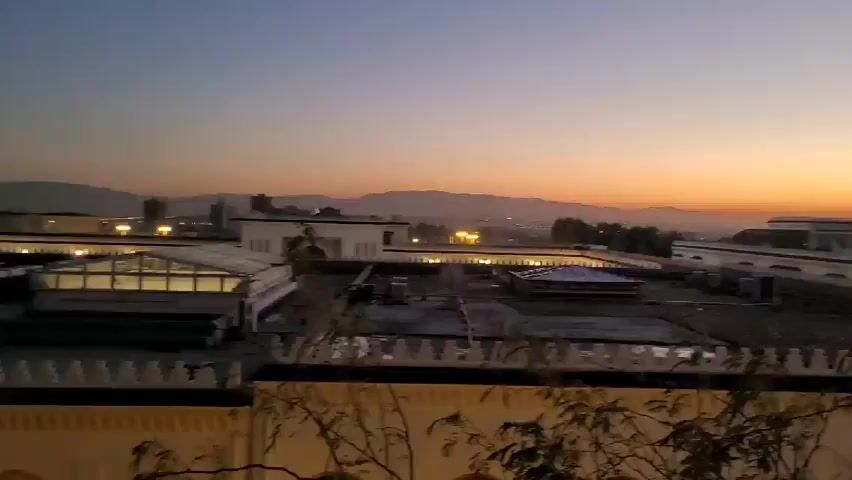 The video captures an elevated view of a cityscape at dawn or dusk. The sky transitions from a deep blue at the top to a vibrant orange near the horizon where silhouettes of mountains are visible. Several buildings are visible, most of which are white or light-colored with distinct architectural details and rooflines. Some buildings have lights illuminating them.  A foreground wall with a decorative top runs along the bottom of the frame. Sparse foliage partially obscures the lower portion of the wall. The camera remains static throughout the short video.