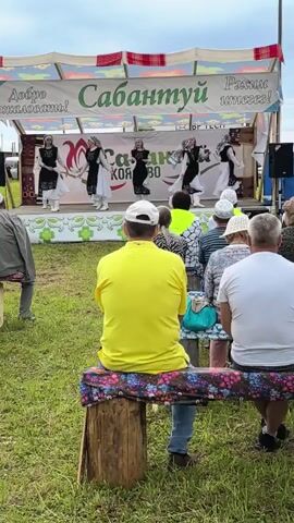 The video shows an outdoor performance on a stage decorated with a banner that reads "Сабантуй" (Sabantuy). Several individuals are performing on stage, dressed in traditional attire. They appear to be dancing or singing. In front of the stage, an audience is seated on benches, watching the performance. The environment is grassy, suggesting a park or open field. The weather appears to be overcast. The focus is on the performance and the audience's engagement.