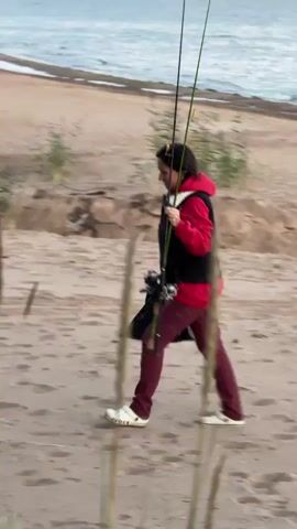 A person wearing a red hooded jacket, a black vest, and burgundy pants is walking on a sandy beach. They are carrying multiple fishing rods and a black bag. The person is looking towards the camera and smiling. In the background, there is a body of water, a sandy shore, and trees. The scene appears to be a bright, outdoor environment.