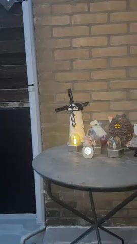 The video shows a close-up view of a round, weathered metal table against a brick wall. On the table are several decorative garden ornaments. From left to right, there is a white windmill with black blades that is lit with a warm yellow light. Next to it is a small figurine of a tan animal, possibly a squirrel or a chipmunk, sitting on a decorative clock or solar light. Behind the animal figurine is a clear jar containing various items, including what appear to be small purple and white objects, possibly candies or decorative stones. To the right of the jar is a miniature house shaped like a pinecone with a red-capped mushroom on top. Further to the right, a part of another small decorative house or lantern is visible. The lighting suggests it is dusk or early evening. The brick wall behind the table is visible in detail.