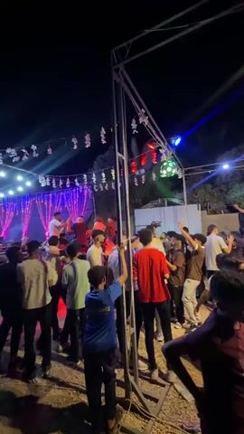 This is a wide shot of an outdoor nighttime gathering. A stage with purple and white lights is set up in the background, and a band appears to be performing. Numerous young people are gathered in front of the stage, some dancing and others observing. A metal structure with lights is prominently placed in the foreground, obstructing part of the view of the crowd. The ground appears to be dirt and gravel, and trees are visible behind the stage area.