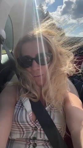 A blonde woman with curly hair, wearing sunglasses and a plaid tank top, is sitting in the passenger seat of a convertible car. The top is down, and her hair is blowing in the wind. The sun is shining brightly, casting a glare on the scene. The road ahead is visible, with a few cars in the distance. The woman smiles at the camera, and at one point, she raises her right arm and touches her hair.