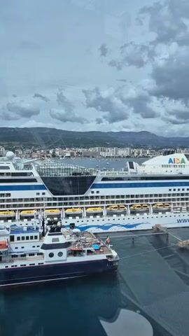 The camera pans upward to reveal a view from inside a cruise ship, through a window overlooking a harbor. Two cruise ships are docked, one labeled “AIDASTELLA”. A small, dark-hulled boat is also docked. In the background, there is a city and a mountain range under a cloudy sky. The phrase "Too lazy to leave the ship" appears on the screen.