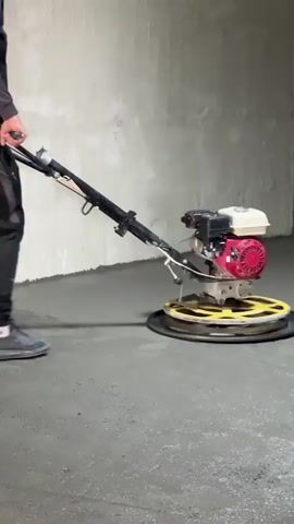 A person operates a concrete power trowel in a bare room. The person, dressed in a long-sleeved black shirt and black pants, guides the trowel across a wet concrete floor. The trowel is a walk-behind machine with a black handle and a red and white engine mounted above a yellow safety cage, enclosing rotating blades. The machine smooths the concrete as it moves. The room has unfinished concrete walls and an open doorway on the right.