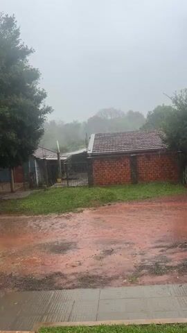 This media file captures a rainy day scene in Encarnacion, Paraguay. The primary subjects are a residential property with a red-tiled roof and a brick wall, and a dirt/gravel driveway or yard in the foreground. The setting appears to be a rural or suburban area, with trees and vegetation surrounding the property. The sky is overcast and a heavy mist or rain is visible, obscuring the distant landscape.

The activity taking place is a rain shower. Water is visibly falling from the sky and pooling on the ground, creating a wet and somewhat dreary atmosphere. The trees and grass appear lush, suggesting this is a place where rain is common.

Notable details include the reddish-brown color of the wet ground, likely due to the soil composition. The brick wall of the property is prominent, adding a rustic element to the scene. The time of day is difficult to determine precisely due to the overcast conditions, but it appears to be daytime. The overall mood is one of tranquility, perhaps even a bit somber, due to the persistent rain. There is no visible text in the media file.