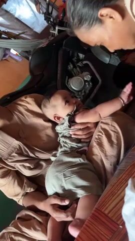 A close-up shot shows a baby being held by a person who is leaning over the baby. The baby is wearing a light-colored onesie and is lying on its back. The person holding the baby has dark hair and is wearing a light-colored top with a pattern. The background is blurred, but it appears to be an indoor setting with some objects that look like part of a vehicle. The lighting is soft and natural. The interaction appears to be tender and caring, with the person looking intently at the baby.