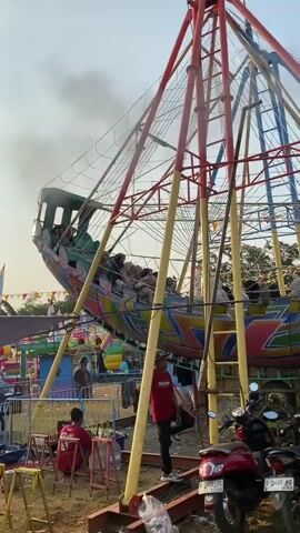 A ride that resembles a Viking ship on a pendulum swing is in operation at an outdoor fair. People are seated inside the ride, which is currently at the apex of its swing. Below the ride, a vendor stall is set up with various drinks and snacks displayed. Several motorcycles are parked in the foreground. The ground is dirt. The sky is clear and bright.