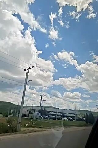 The video shows a view from inside a vehicle, looking out at a bright, cloudy sky and a street scene. On the right side of the frame, a silver car is partially visible, driving on the road. In the background, there is a long, low-rise building with a white facade and a dark roof, possibly a commercial establishment. Several cars are parked in front of this building. Power lines and utility poles are prominent in the sky. The overall impression is of a suburban or industrial area on a sunny day. The camera is moving, indicating the footage was taken while in transit.