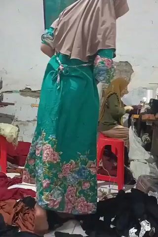THE VIDEO SHOWS A WOMAN WEARING A TEAL DRESS WITH FLORAL PATTERNS AND A TAN HIJAB STANDING IN A ROOM. ANOTHER WOMAN IS SEEN IN THE BACKGROUND, WEARING A GREEN OUTFIT AND A YELLOW HIJAB, SITTING ON A RED STOOL AND OPERATING A SEWING MACHINE. THE ROOM APPEARS TO BE A WORKSHOP OR A TAILORING STUDIO, WITH VARIOUS FABRICS AND SEWING EQUIPMENT VISIBLE. THE LIGHTING IS DIM AND NATURAL.