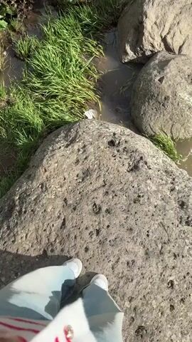 The video shows a view of a stream or small river. The camera angle is from a person standing on a large gray rock, looking down at the water and surrounding rocks. The water is murky and flowing, with visible rocks and grassy areas on the banks. There is greenery present, with green grass growing amongst the rocks near the water. The person is wearing light-colored jeans and light-colored sneakers, and the bottom of their pants is visible. The video is taken during the daytime.