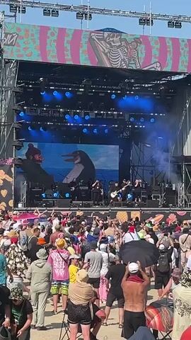 A wide shot shows a large outdoor music festival stage with a band performing. A large screen behind the band displays animated characters, a penguin and a person. The stage is equipped with lights and speakers. In front of the stage, a large crowd of people are gathered, many holding up their phones to record. The environment appears to be a beach or open area with a clear blue sky and some water visible in the background. The crowd is diverse in age and attire, with many wearing summer clothing. The overall atmosphere appears to be energetic and festive.