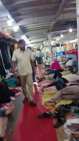 The camera pans down from a high ceiling in a large room to a scene where numerous people are lying down on blankets and what appear to be thin mattresses, occupying much of the visible floor space. The room has an industrial feel, indicated by the ceiling structure with prominent supports and industrial lighting fixtures. Several people are seen lying relatively still, some appearing to be sleeping or resting, while others are sitting or in semi-reclined positions. The blankets and mattresses are colorful and patterned. The overall impression is of a makeshift or temporary sleeping area in a large, possibly repurposed, indoor space.