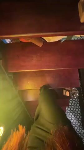 The video begins with a first-person view of descending a dark wooden staircase. The camera pans slightly, revealing patterned flooring and a green wall with various objects on shelves. As the descent continues, the view shifts to a dimly lit bar area. A person wearing an apron and jeans is visible standing behind the bar, which is lined with numerous bottles and glasses. The bar itself is green, and a wine cooler is situated beneath the shelves on the left side of the frame. The ceiling has a grid-like structure with ceiling fans.