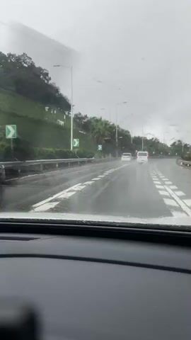 The video shows a view from inside a car driving on a wet road in the rain. The camera is focused on the road ahead, with a black SUV in the left lane and other vehicles further down the road. The scenery includes a grassy hill on the left side and palm trees lining the road. The dashboard of the car is partially visible in the foreground, showing a digital display. The road appears to be a highway or a major road with multiple lanes. The weather is overcast and rainy, with water splashing from the tires of the vehicles. The black SUV has distinctive red taillights and appears to be moving at a moderate speed. The video captures the movement of the vehicles and the environmental conditions of a rainy day drive.