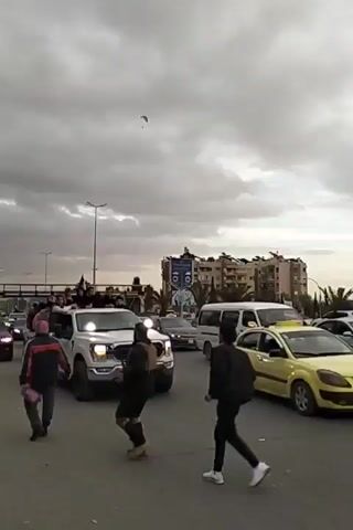 The video shows a street scene in what appears to be an urban area. A white pickup truck is driving through the intersection carrying approximately a dozen people in its bed and along the cab's roof. Several individuals are standing near the vehicle. Other cars, including a police car, a white van, and a yellow taxi, are visible in the surrounding traffic. Overhead, a single paraglider is visible against a cloudy sky. Buildings, streetlights, and an overpass are also present in the background. The overall atmosphere suggests a gathering or celebration.