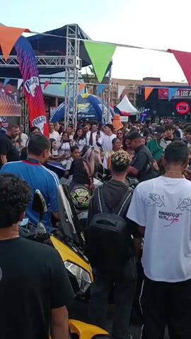 The video captures a scene from a crowded outdoor event, possibly a motorcycle rally or festival. A diverse crowd of people, including adults and children, are gathered around several yellow motorcycles. Many are holding up phones, seemingly taking photos or videos of something of interest. There is a stage in the background with banners and signs, suggesting that the event is sponsored or organized. Several people in the crowd are wearing casual clothes like t-shirts and jeans. The atmosphere is lively, with visible excitement among the attendees. The environment is open-air, with visible buildings and decorations contributing to the event's festive atmosphere.