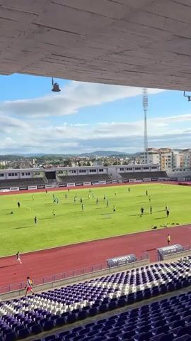 A soccer practice is underway in a stadium. Several players in blue and purple uniforms are scattered across a bright green field, some dribbling soccer balls, others running. A red running track surrounds the field, and beyond that, purple and white stadium seats are visible. In the background, a town with buildings and mountains stretches under a partly cloudy blue sky. A tall light tower stands on the right side of the frame. The camera is positioned under a white overhang, looking down and across the field.