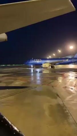 The video shows a blue ITA Airways airplane parked on a wet tarmac at night. Streetlights illuminate the scene. The footage appears to be taken from the window of another airplane, with the wing visible in the foreground. The tarmac is marked with red and white lines.