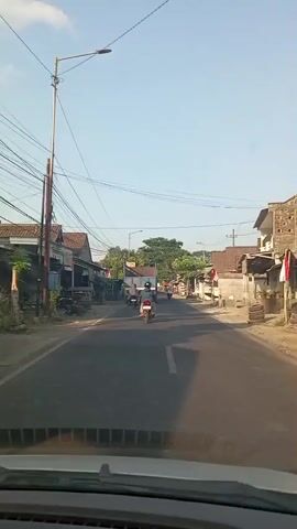The video shows a view from inside a vehicle, looking forward down a paved road in a village. The road is lined with small buildings, some with shops or houses. There are utility poles and overhead wires along the side of the road. A few motorcycles and a truck are visible on the road. Trees and vegetation are present between the buildings and further down the road. The sky is clear and blue, indicating daylight. The overall impression is of a typical rural or semi-urban street scene.