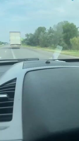 This media file depicts a view from inside a vehicle, looking forward through the windshield. The primary subjects are the road ahead, a large semi-trailer truck in the distance, and a billboard. The scene is set on a highway in Ukraine, specifically near Pidhorodnie, as indicated by the location context.

The activity taking place is driving on a highway. The truck is in the left lane, and the viewpoint vehicle is in the right lane, suggesting overtaking or a similar maneuver is possible. The billboard is positioned to the right of the road and is advertising a product or service. The weather appears to be overcast with a bright sky, indicating it is daytime.

Notable details include the dashboard of the vehicle in the foreground, with vents and a small circular object, possibly a sensor. The truck has a white trailer with some green lettering. The billboard has yellow and green coloring and features an image of a character holding fruit. Visible text on the billboard, though partially obscured and blurry, appears to be in Ukrainian. It reads "ХАПАН СВІН," which translates to "Swine Capture" or "Pig Catch," possibly related to a farming or agricultural product. The overall mood conveyed is one of travel and observation from a moving vehicle.