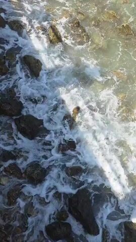 The video shows a close-up, high-angle view of waves crashing against rocks on a shore. The water is clear and shallow, with sunlight reflecting off the surface and illuminating the rocks beneath. The rocks are of various sizes and shapes, some smooth and rounded, others rough and jagged. The waves create white foam and churn the water as they break against the shore. The sound of the waves is audible. The scene is natural and peaceful.
