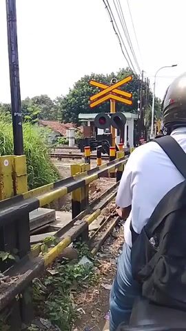The video shows a POV of someone riding a motorcycle, approaching a railway crossing. A red and white railway signal is visible, along with a yellow and black striped barrier. Several other motorcycles and a white car are stopped at the crossing, waiting. The person riding the motorcycle is wearing a white shirt and a helmet, with a black backpack. Trees and buildings line the background. The railway crossing signal changes to red, indicating an approaching train. One person on a motorcycle in front is wearing a red jacket and beige pants. Another person is on a motorcycle with a green basket attached. The video captures the moment of waiting at the railway crossing as traffic is halted.