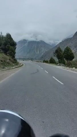 A first-person perspective video shows a paved road winding through a mountainous valley under a cloudy sky. The road is gray asphalt with white lane markings. To the left of the road, a steep, rocky embankment is covered with sparse vegetation and some trees. To the right, the terrain slopes more gently, with trees and a distant view of more mountains. The mountains in the background are large and imposing, with some peaks obscured by thick clouds. The overall impression is one of a remote and scenic landscape, likely a mountain pass. The bottom of the frame shows a portion of a dark helmet and handlebar, suggesting the video is being recorded from a motorcycle or bicycle.